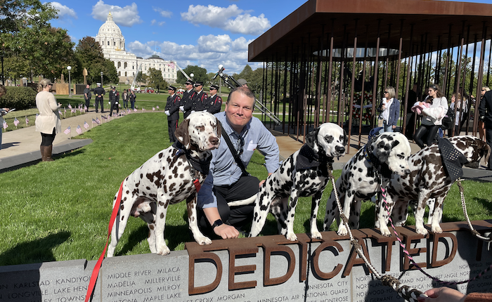 A man poses with 4 Dalmatian dogs behind a sign that reads Dedicated